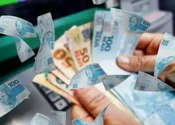 Mãos segurando cédulas de R$ 50 e R$ 100 em frente a um caixa eletrônico, com notas voando ao redor em composição dinâmica, ilustrando a liberação automática das parcelas extras do Seguro-Desemprego para trabalhadores afetados pelas chuvas em Minas Gerais