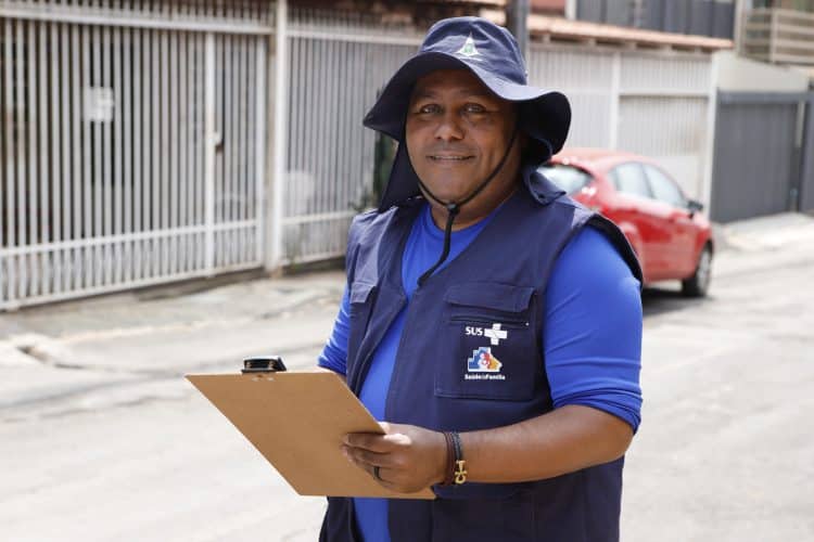 Agente comunitário do SUS sorri segurando prancheta em rua residencial