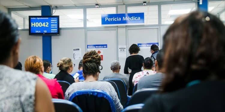 Sala de espera de agência do INSS com pessoas aguardando atendimento para perícia médica e painel eletrônico exibindo senhas representando a mudança no sistema de atendimento para reduzir filas