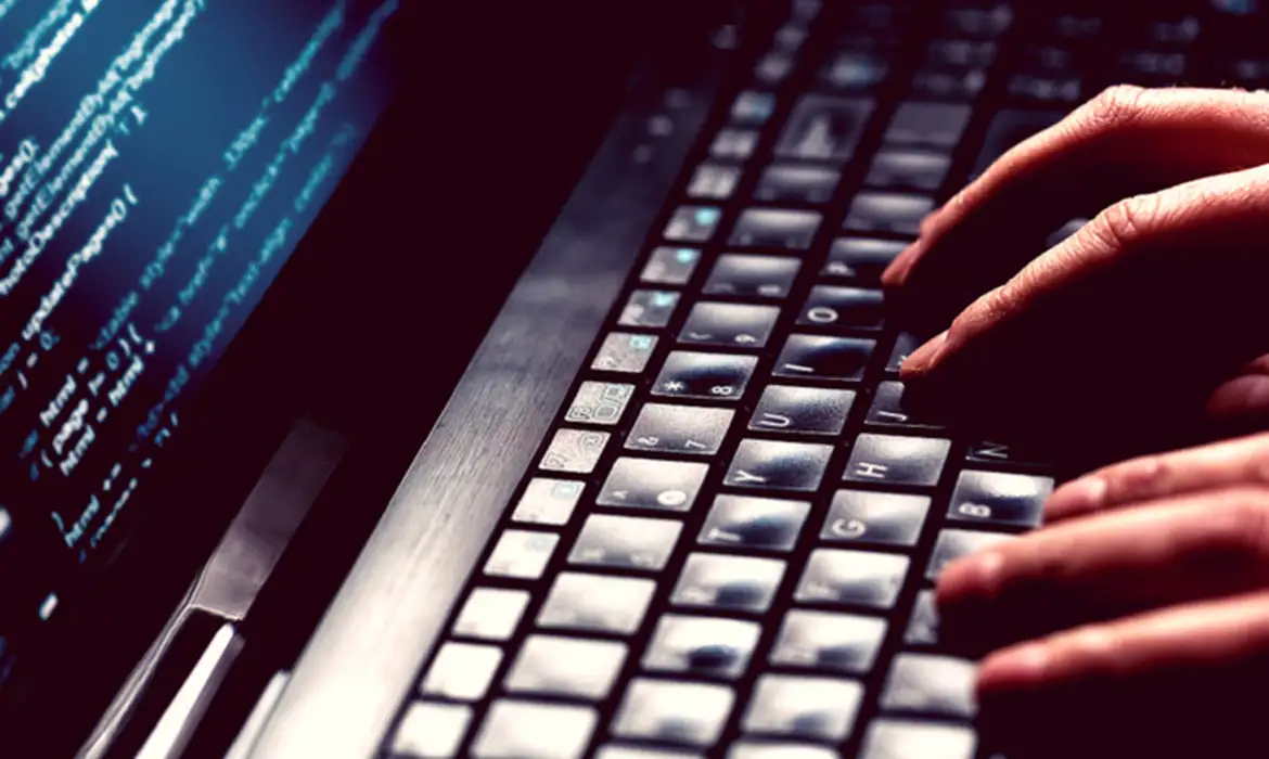 Mãos digitando em teclado de notebook com código de programação na tela em ambiente escuro, representando as aulas práticas de cibersegurança do programa Hackers do Bem
