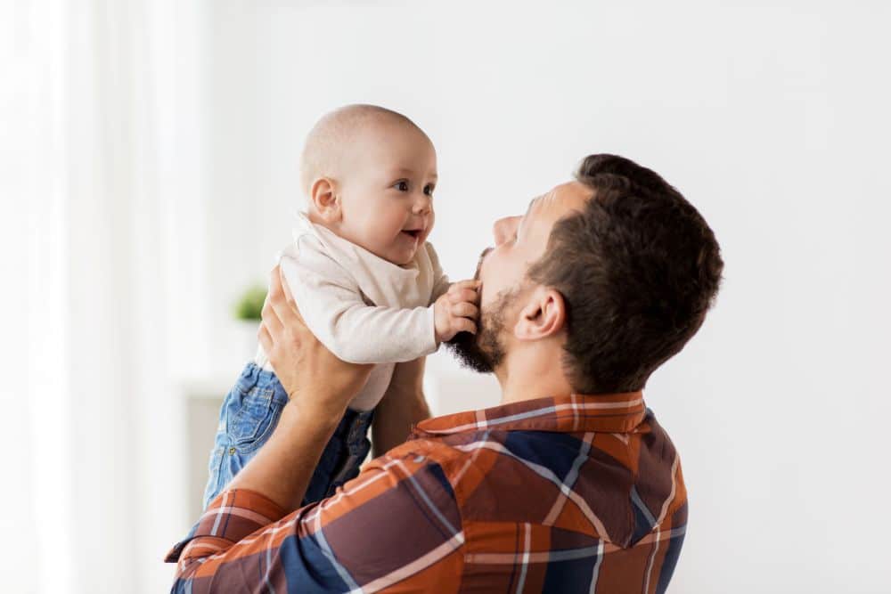 Um homem barbudo vestindo uma camisa xadrez laranja e azul segura um bebê careca no alto. O bebê está de body branco e calça jeans, e os dois se olham sorrindo com carinho, em um ambiente interno bem iluminado.