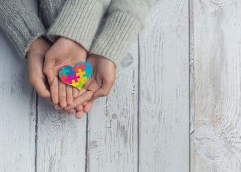 Close das mãos de uma criança e de um adulto sobre uma mesa de madeira clara, segurando juntos um pequeno coração colorido com estampa de peças de quebra-cabeça, símbolo do Transtorno do Espectro Autista (TEA).