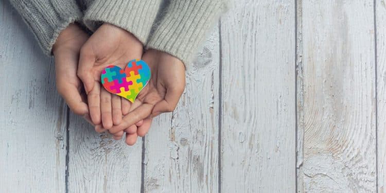 Close das mãos de uma criança e de um adulto sobre uma mesa de madeira clara, segurando juntos um pequeno coração colorido com estampa de peças de quebra-cabeça, símbolo do Transtorno do Espectro Autista (TEA).