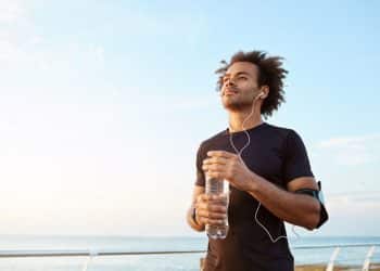 Close de um homem jovem e atlético, de pele negra e cabelo afro, vestindo roupa preta de corrida, com fones de ouvido e uma garrafa de água na mão, olhando pensativo para o horizonte ao pôr do sol em um calçadão à beira-mar.