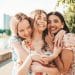 Três amigas sorridentes se abraçando ao ar livre em um dia ensolarado, transmitindo afeto e bem-estar.