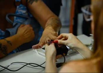 Close-up das mãos de uma pessoa realizando a coleta de impressão digital em um scanner biométrico sobre uma mesa branca. Uma atendente auxilia no posicionamento do dedo sobre o sensor.