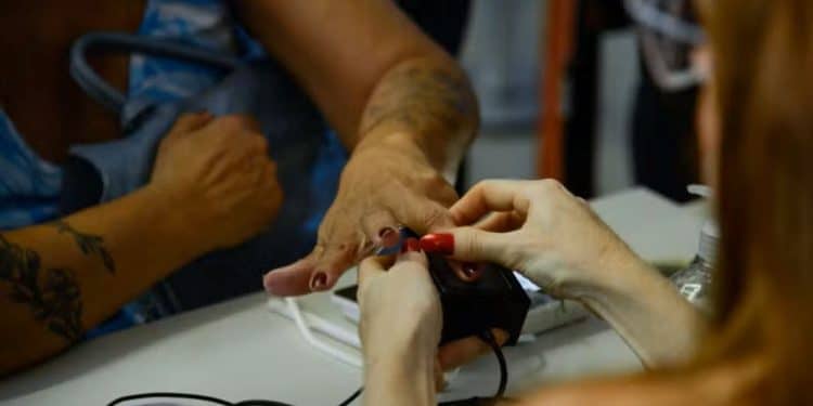 Close-up das mãos de uma pessoa realizando a coleta de impressão digital em um scanner biométrico sobre uma mesa branca. Uma atendente auxilia no posicionamento do dedo sobre o sensor.