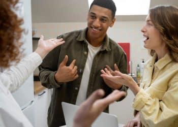 Três jovens em uma cozinha conversando de forma animada; um rapaz negro ao centro sorri enquanto duas mulheres gesticulam positivamente durante o diálogo.