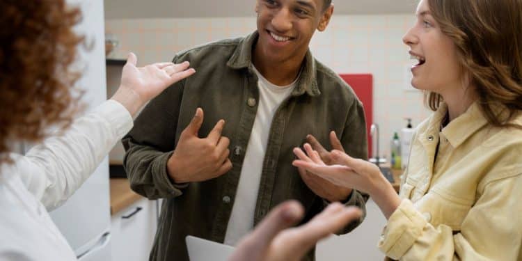 Três jovens em uma cozinha conversando de forma animada; um rapaz negro ao centro sorri enquanto duas mulheres gesticulam positivamente durante o diálogo.