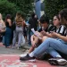 Grupo de estudantes sentados na calçada consultando papéis e celular enquanto outros aguardam em fila na entrada de uma instituição de ensino.