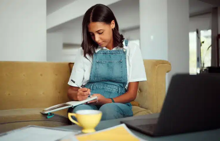 Jovem sentada em sofá anotando em caderno com notebook e xícara amarela à frente