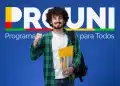 Estudante jovem com mochila e cadernos sorrindo à frente de um painel azul com o logotipo do Prouni (Programa Universidade para Todos). Ele faz um gesto de celebração com o punho cerrado.