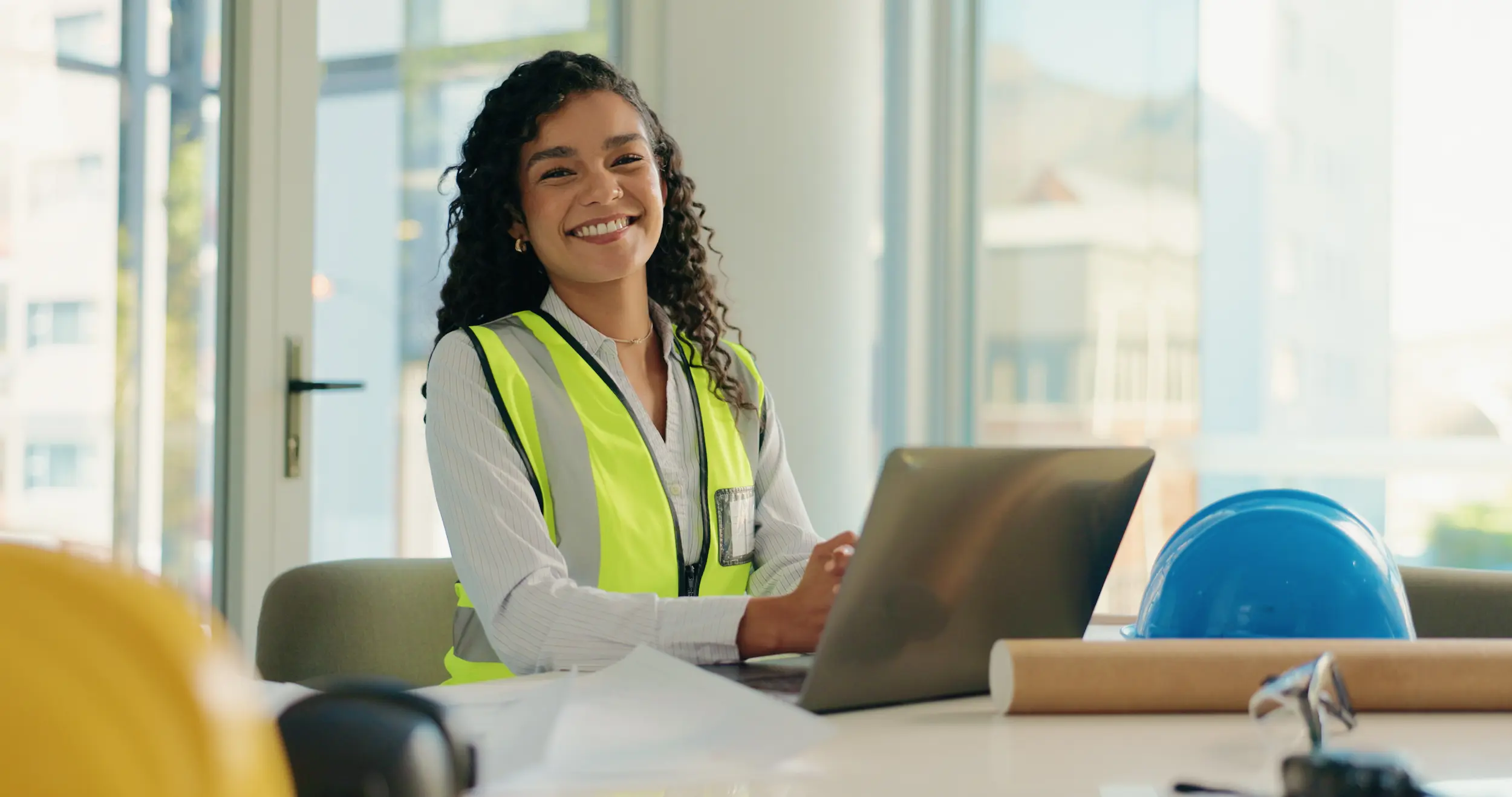 Engenheira sorrindo enquanto trabalha no notebook em ambiente de obra ou projeto, representando os desafios enfrentados pelas mulheres para crescer profissionalmente.