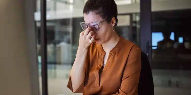 Mulher com dor de cabeça, claramente sobrecarregada de trabalho, em escritório com documentos em cima da mesa, expressão de frustração e cansaço.