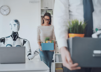 Robô humanoide trabalhando em notebook enquanto funcionários humanos carregam caixas de pertences ao sair do escritório