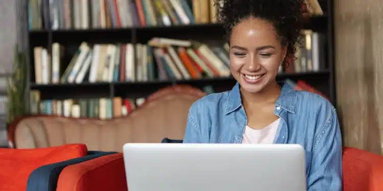 Estudante sorrindo ao usar notebook em biblioteca, acessando portal de certificados