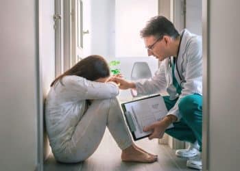 Médico acolhendo mulher que chora sentada no chão, demonstrando empatia e cuidado com a saúde emocional