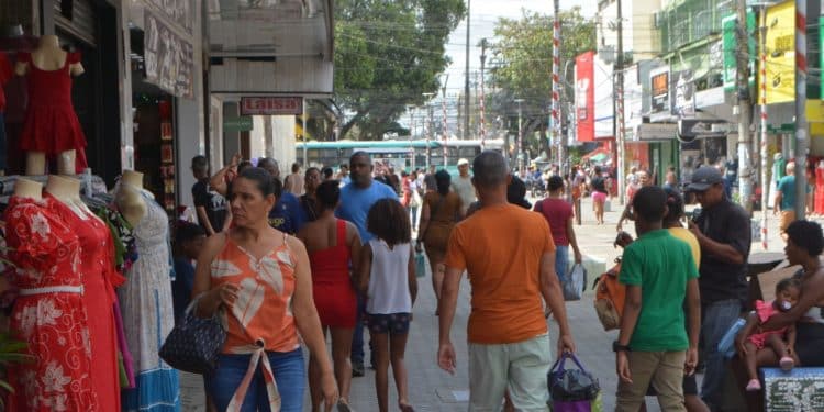Pessoas circulam entre vitrines e manequins com vestidos coloridos em rua movimentada de comércio.