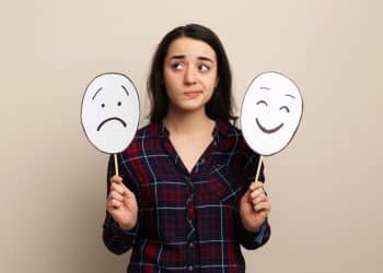 Jovem segurando duas máscaras com expressões de tristeza e felicidade simbolizando o controle das emoções.