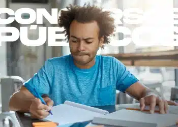 Homem jovem de cabelo afro e camiseta azul estudando concentrado com cadernos e canetas, com a sobreposição de texto "Concursos Públicos".