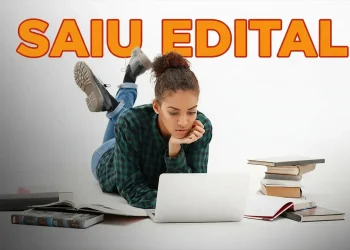 Jovem estudando no computador enquanto lê livros, anunciando novo concurso com 80 vagas e salários de até R$ 9,6 mil.
