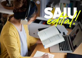 Candidata estudando com fone de ouvido e livro aberto ao lado de notebook com texto Saiu Edital — concurso público Goianinha RN salários até R$ 6,1 mil 2026