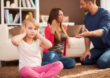 Menina cobre os ouvidos enquanto presencia um desentendimento entre os pais na sala de casa.