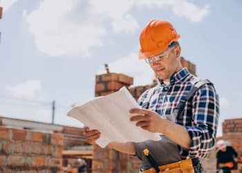 Jovem trabalhador da construção civil em um canteiro de obras ao ar livre, vestindo capacete laranja, óculos de segurança e camisa xadrez, concentrado na leitura de uma planta técnica de papel.