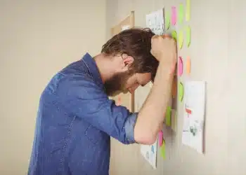 Homem frustrado encostando a cabeça em um mural com post-its coloridos, simbolizando dificuldade em organizar rotinas e controlar impulsos.