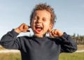 Imagem frontal de uma criança com cabelo cacheado e uma camisa azul marinho, com os olhos fechados, a boca aberta gritando e os dedos indicadores tapando os ouvidos.