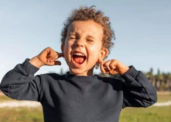 Imagem frontal de uma criança com cabelo cacheado e uma camisa azul-marinho, com os olhos fechados, a boca aberta gritando e os dedos indicadores tapando os ouvidos.