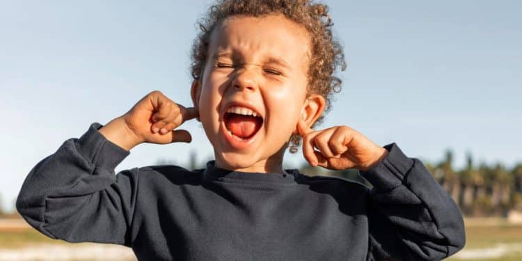 Imagem frontal de uma criança com cabelo cacheado e uma camisa azul-marinho, com os olhos fechados, a boca aberta gritando e os dedos indicadores tapando os ouvidos.