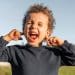 Imagem frontal de uma criança com cabelo cacheado e uma camisa azul-marinho, com os olhos fechados, a boca aberta gritando e os dedos indicadores tapando os ouvidos.