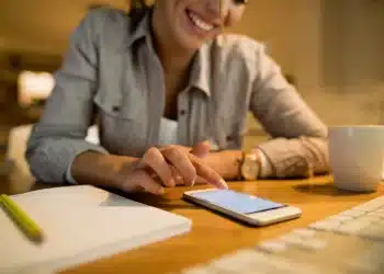 ulher sorrindo toca a tela do celular enquanto prepara currículo ao lado de caderno