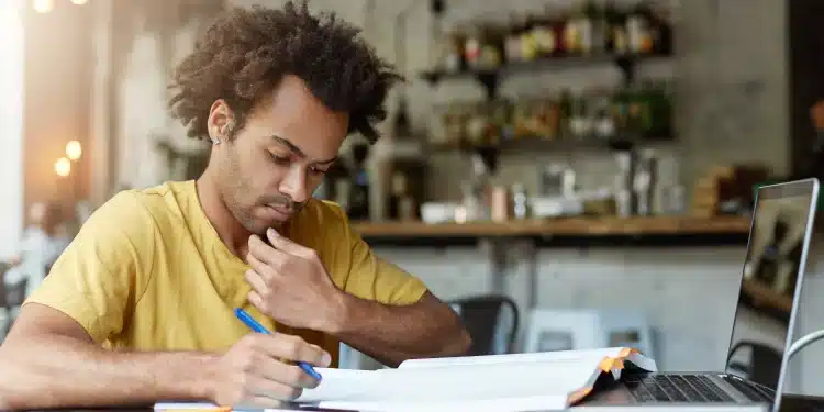 Jovem escreve em caderno enquanto consulta livro aberto ao lado de notebook em mesa