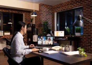 Mulher participando de videoconferência pelo notebook em escritório doméstico com parede de tijolos ilustrando a rotina de trabalho remoto