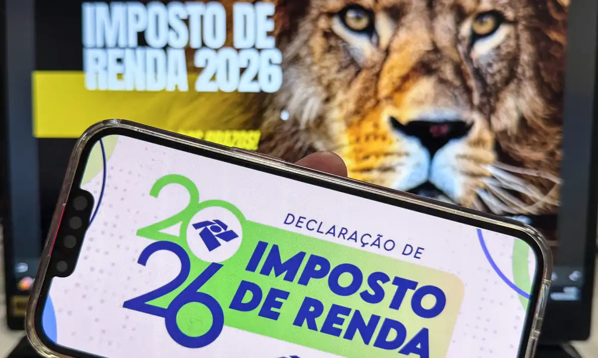 Mão segurando celular com o aplicativo da Declaração de Imposto de Renda 2026 na tela, com imagem do Leão da Receita Federal ao fundo, simbolizando o início do prazo de entrega da declaração e as novidades do IRPF 2026
