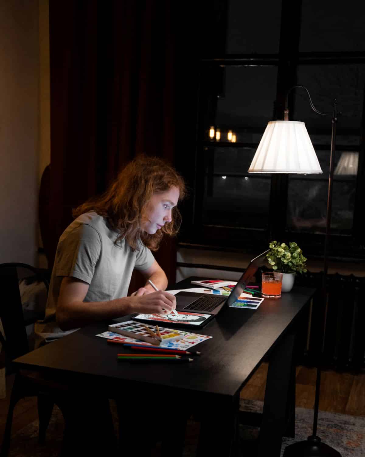 Um jovem de cabelos longos e ondulados trabalha concentrado em uma mesa escura à noite. Ele utiliza uma caneta digital em um tablet gráfico conectado a um notebook, cercado por aquarelas, lápis de cor e paletas de cores, sob a luz de uma luminária de pé.