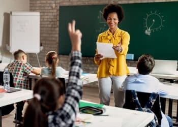 Professora negra sorridente em sala de aula, segurando um papel e interagindo com alunos sentados em suas bancas, em frente a um quadro verde.