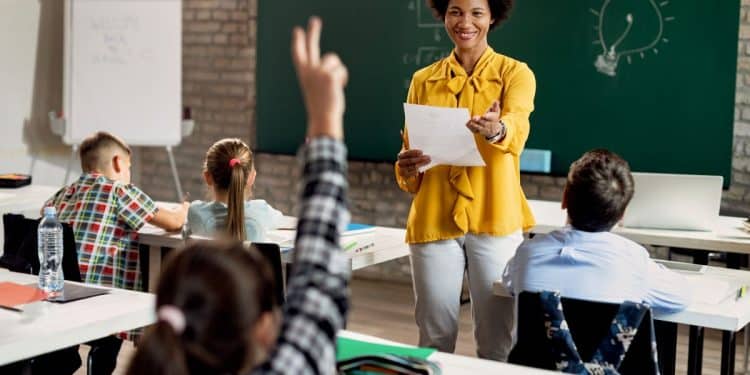 Professora negra sorridente em sala de aula, segurando um papel e interagindo com alunos sentados em suas bancas, em frente a um quadro verde.