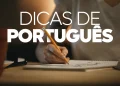 Close-up das mãos de uma pessoa escrevendo em um caderno com um lápis. Ao fundo, um vaso de planta desfocado. Sobre a imagem, o texto em destaque diz "DICAS DE PORTUGUÊS". A iluminação é suave e focada na área de estudo.