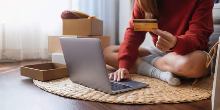 Mulher sentada no chão usando notebook e cartão de crédito rodeada de caixas de encomendas