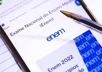 Close-up de um smartphone exibindo o logotipo oficial do Enem na tela, com uma caneta esferográfica apoiada sobre o dispositivo, simbolizando a rotina de estudos digital e prática.