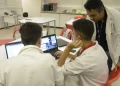 Dois jovens estagiários trabalhando em laptops em uma sala, com um supervisor de jaleco branco orientando, representando o ambiente de estágio profissional e os direitos trabalhistas propostos pelo PL 6350/25