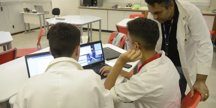Dois jovens estagiários trabalhando em laptops em uma sala, com um supervisor de jaleco branco orientando, representando o ambiente de estágio profissional e os direitos trabalhistas propostos pelo PL 6350/25.