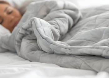 Mulher dormindo profundamente em cama branca, totalmente envolvida por um edredom cinza espesso que cobre até os ombros.