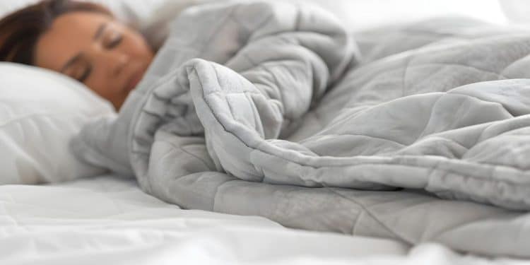 Mulher dormindo profundamente em cama branca, totalmente envolvida por um edredom cinza espesso que cobre até os ombros.