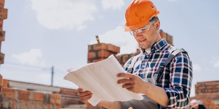 Homem com capacete laranja e óculos de proteção analisando uma planta de construção em um canteiro de obras ao ar livre.