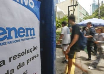 Candidatos caminhando em frente a local de aplicação do Enem com banner azul e branco do Exame Nacional do Ensino Médio em primeiro plano com os dizeres sobre a resolução da prova, em área externa com árvores e prédios ao fundo