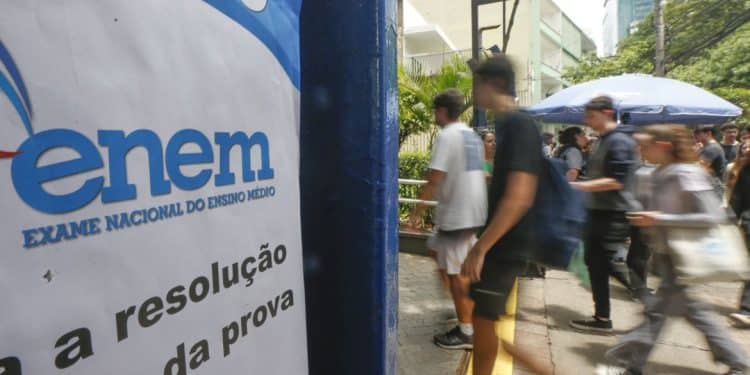 Candidatos caminhando em frente a local de aplicação do Enem com banner azul e branco do Exame Nacional do Ensino Médio em primeiro plano com os dizeres sobre a resolução da prova, em área externa com árvores e prédios ao fundo